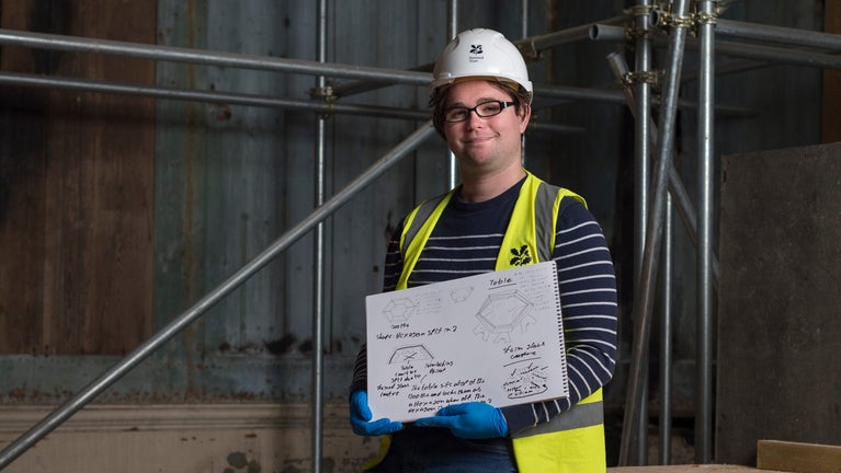 James Naylor wears PPE and faces the camera holding a sketch book with line drawing designs